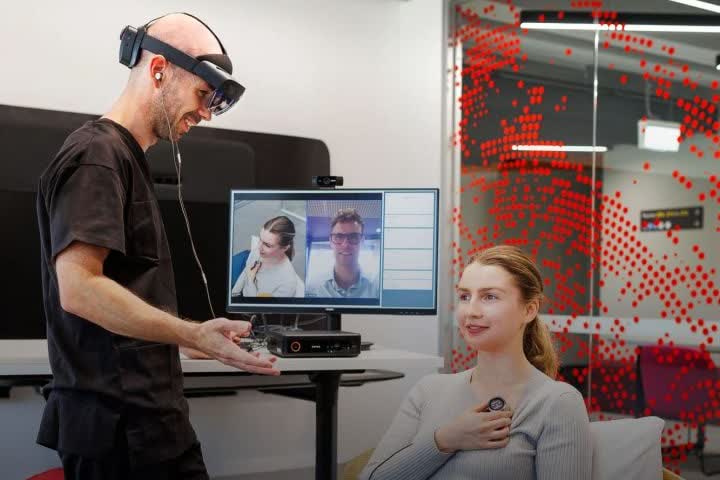 A man in scrubs wearing a HoloLens 2 stands beside a seated woman who holds a stethoscope to her chest; behind them a monitor shows two people on a video call beside a panel of readings