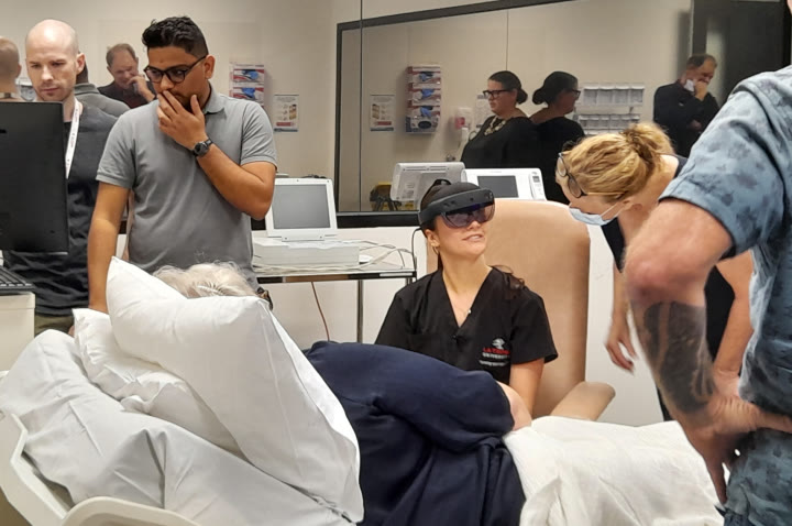 A doctor wearing a HoloLens 2 sits beside a stand-in patient in a hospital bed, while a group of people watch from around the bed